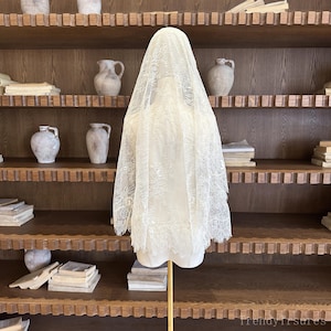 Op de afbeelding: Een cr&egrave;mekleurige kanten sluier, tentoongesteld op een mannequin. De sluier heeft een delicaat bloemmotief en een geschulpte rand. De mannequin staat op een gouden standaard, tegen een achtergrond van houten planken met aardewerk en boeken.