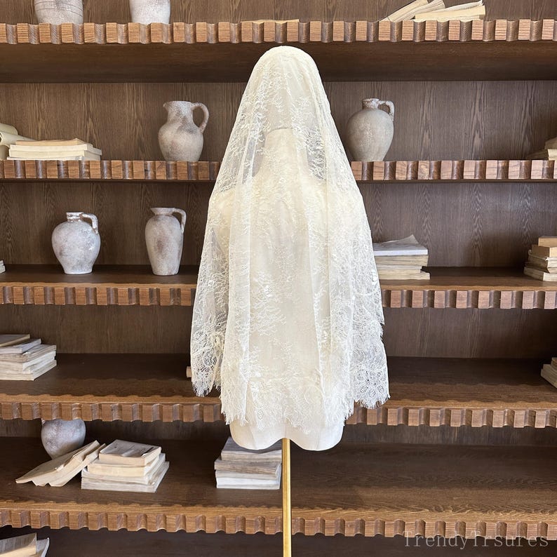 Op de afbeelding: Een cr&egrave;mekleurige kanten sluier, tentoongesteld op een mannequin. De sluier heeft een delicaat bloemmotief en een geschulpte rand. De mannequin staat op een gouden standaard, tegen een achtergrond van houten planken met aardewerk en boeken.