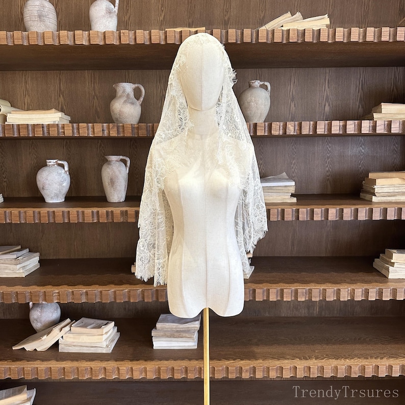 Op de afbeelding: Een cr&egrave;mekleurige kanten sluier, met gedetailleerde bloempatronen en randen, is over een mannequin gedrapeerd. De mannequin staat op een gouden standaard. De achtergrond bestaat uit houten planken met decoratieve items.
