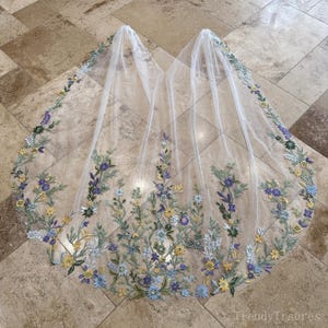 Könnte beinhalten: Ein durchsichtiger weißer Brautschleier mit einem floralen, gestickten Rand. Die Stickerei zeigt Blumen in Blau, Gelb und Grün. Der Schleier liegt auf einer gefliesten Oberfläche.