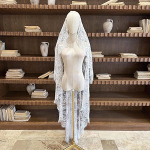 May include: A white lace bridal veil is displayed on a mannequin. The veil is long, with intricate floral lace detailing. The mannequin is on a gold stand, and the background features a wooden bookshelf with books and decorative vases.