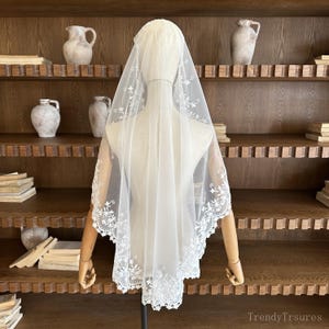 May include: A white bridal veil with delicate floral lace trim. The veil is displayed on a mannequin, showcasing its sheer fabric and intricate embroidered details. A classic accessory for weddings and special occasions.