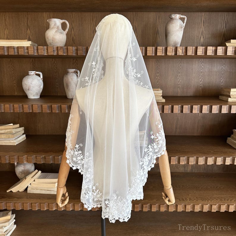 May include: A white bridal veil with delicate floral lace trim. The veil is displayed on a mannequin, showcasing its sheer fabric and intricate embroidered details. A classic accessory for weddings and special occasions.