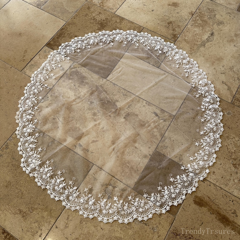 May include: A circular, sheer white veil with a delicate floral lace trim. The lace features intricate floral and leaf patterns, creating a romantic and elegant look. The veil is laid out on a neutral-toned surface.
