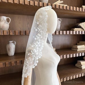 May include: A white bridal veil with a delicate floral lace trim. The veil is draped over a mannequin, showcasing the intricate details of the lace design. The lace trim features a scalloped edge and floral patterns.