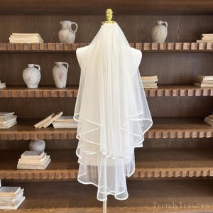 May include: A white bridal veil with a layered design and a delicate trim. The veil is displayed on a mannequin, showcasing its flowing fabric and elegant style. The veil is a classic accessory for weddings.