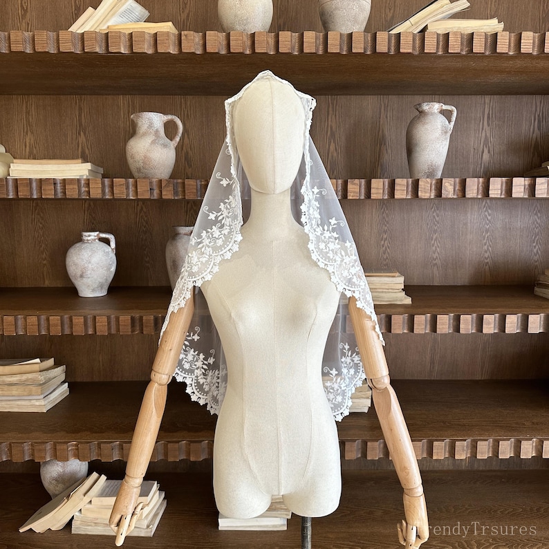 May include: A white lace veil with floral embroidery is displayed on a mannequin. The veil is draped over the mannequin's head and shoulders, showcasing the delicate lace design. The background features a wooden shelf with decorative pottery.
