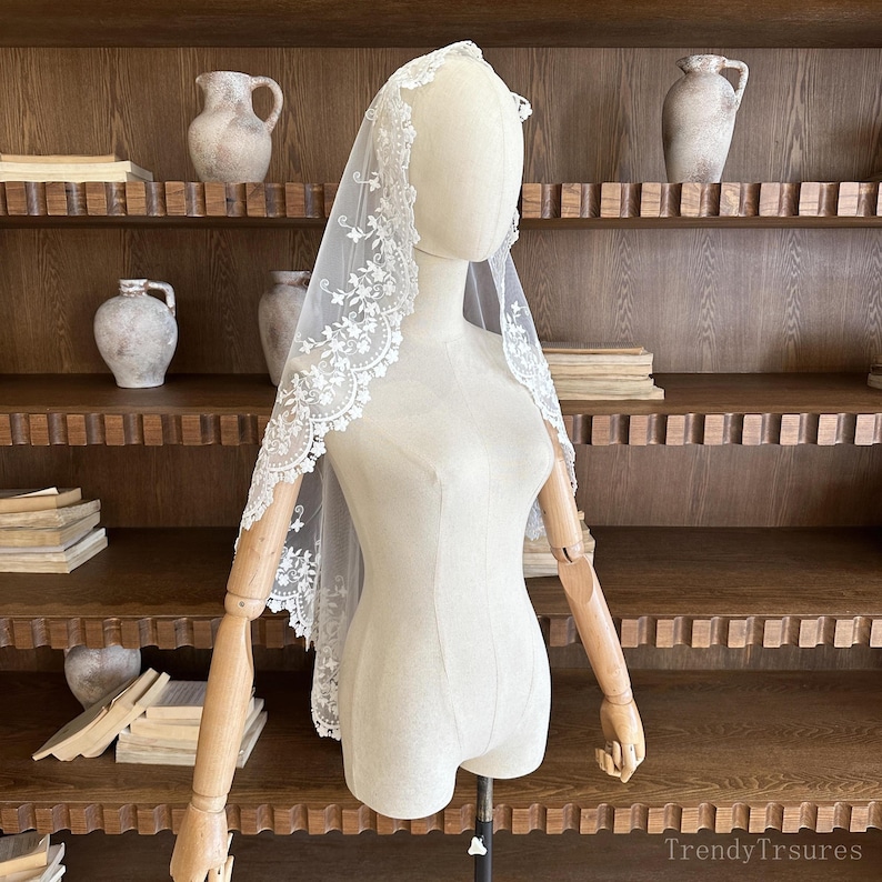 May include: A white bridal veil with delicate floral lace trim is draped over a mannequin. The veil is sheer, allowing the mannequin's form to be visible. The lace trim features intricate floral patterns. The background includes wooden shelves and decorative pottery.