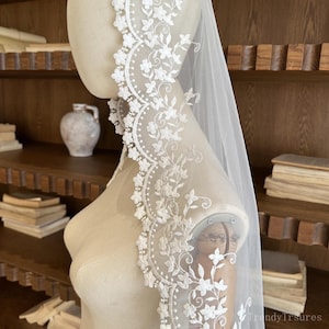 May include: A white bridal veil with a detailed floral lace trim. The lace features intricate flower and leaf patterns. The veil is displayed on a mannequin, showcasing its sheer fabric and design.
