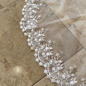May include: Close-up of white lace trim with a floral design. The trim features delicate, embroidered flowers and small pearl-like beads along a scalloped edge. The lace is set against a neutral background.