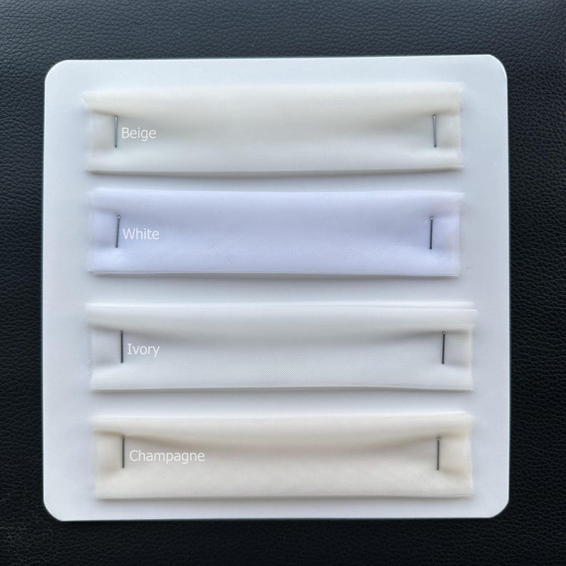 May include: A white card shows four fabric swatches in Beige, White, Ivory, and Champagne. Each swatch is held in place with a silver pin. The colour names are printed above each swatch.