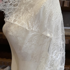 Op de afbeelding: Ivoor kanten stof gedrapeerd over een mannequin. De delicate kant heeft ingewikkelde bloempatronen en een geschulpte rand. De stof is semi-transparant en onthult de vorm eronder. Het algehele effect is elegant en romantisch.