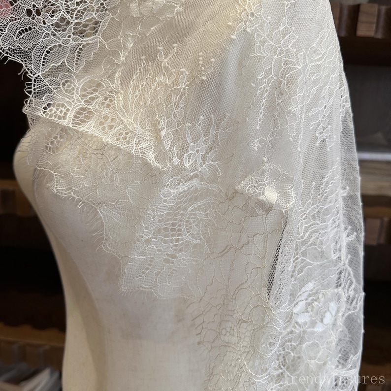 Op de afbeelding: Ivoor kanten stof gedrapeerd over een mannequin. De delicate kant heeft ingewikkelde bloempatronen en een geschulpte rand. De stof is semi-transparant en onthult de vorm eronder. Het algehele effect is elegant en romantisch.