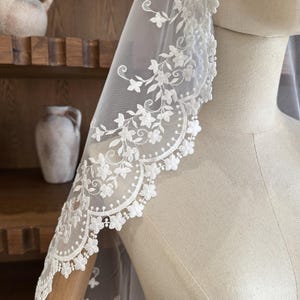 May include: A white bridal veil with a floral lace trim. The lace is adorned with small flowers and leaves, creating an elegant design. The veil is draped over a mannequin, showcasing its sheer fabric and intricate details.