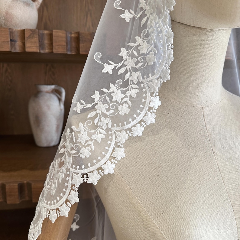 May include: A white bridal veil with a floral lace trim. The lace is adorned with small flowers and leaves, creating an elegant design. The veil is draped over a mannequin, showcasing its sheer fabric and intricate details.