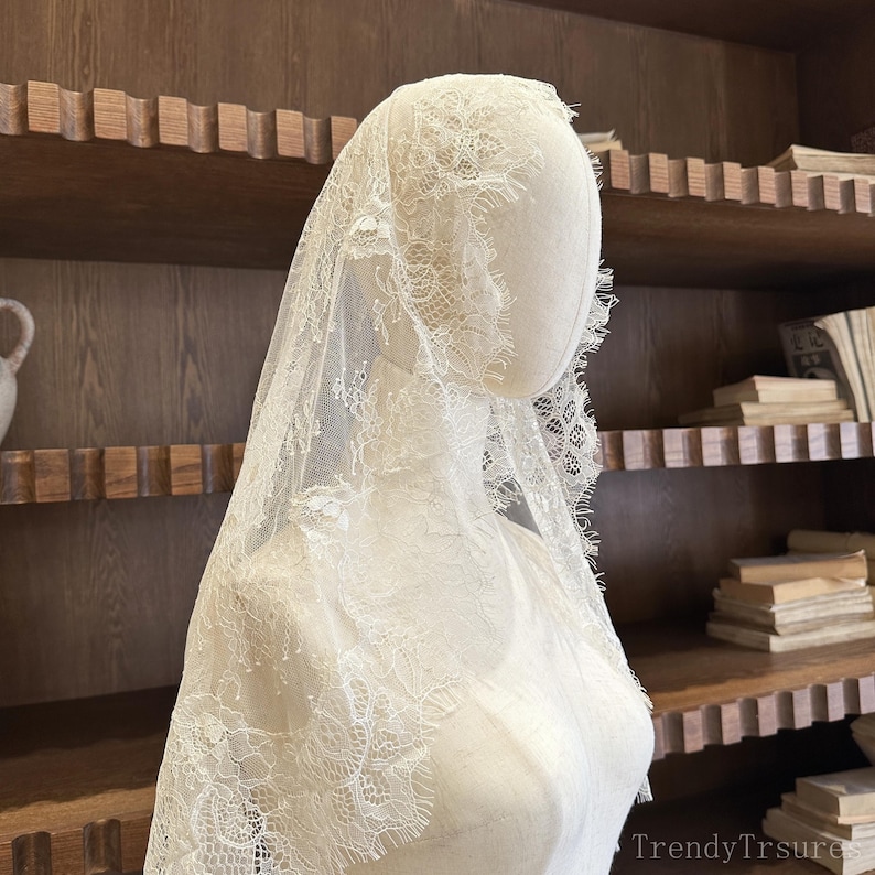 Op de afbeelding: Een witte kanten sluier met ingewikkelde bloempatronen is over een mannequin gedrapeerd. De delicate stof wordt gepresenteerd tegen een achtergrond van een houten boekenplank. De randen van de sluier zijn geschulpt, wat bijdraagt aan het elegante ontwerp. De algehele esthetiek is vintage en romantisch.
