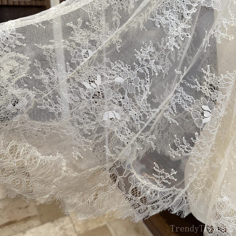 Op de afbeelding: Close-up van delicate witte kanten stof met ingewikkelde bloempatronen. Het kant heeft een geschulpte rand en is licht doorschijnend, waardoor de textuur en het ontwerp zichtbaar worden. De stof wordt waarschijnlijk gebruikt voor kleding of woondecoratie.
