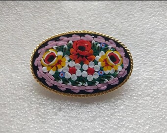 May include: An oval-shaped brooch with a mosaic design featuring colorful flowers on a black background. The brooch is surrounded by a gold-colored metal frame.