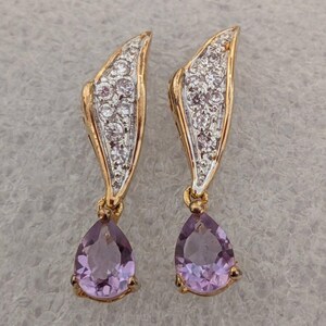 May include: A pair of gold-toned earrings with a leaf-like design. The top portion is encrusted with small, clear stones, while the bottom features a teardrop-shaped, light purple gemstone. The earrings are set against a neutral background.