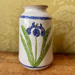 May include: A ceramic vase with a white speckled finish, featuring a blue iris flower design. The vase has a blue band at the top and bottom, with green leaves. The rim is a natural tan color. A decorative piece for the home.