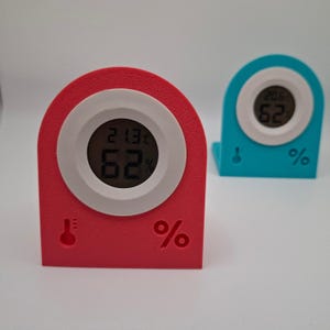 May include: Two digital hygrometer-thermometer devices with arched frames. One is bright pink and the other is teal. Each features a circular white centre with a digital display showing 21.3°C and 62% humidity, plus embossed thermometer and percent icons.