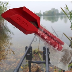 Puede incluir: Un recogedor de cebo de plástico rojo con el texto "Pl Ph" vierte cebo en una bolsa de plástico transparente. El fondo muestra un lago y cañas de pescar, lo que sugiere un producto de pesca.