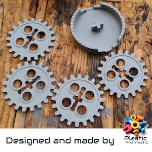 May include: Five grey plastic gears with a centre hole and a grey plastic base with a centre hole. The gears are designed to interlock. The base is designed to hold the gears in place. The text "Designed and made by Plastic Phantastic" is printed below the gears.