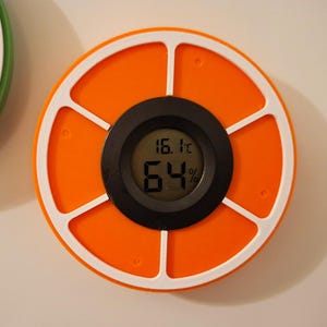 Könnte beinhalten: Ein runder, orangefarbener Digitalthermometer mit weißem, segmentiertem Rahmen. Die schwarze Digitalanzeige zeigt 16,1°C und 64% an. Das Gerät, mit einem schwarzen Mittelteil, misst wahrscheinlich Temperatur und Luftfeuchtigkeit.
