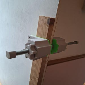 Puede incluir: Una abrazadera blanca y verde está sujeta a un marco de madera. La abrazadera tiene una varilla roscada y una perilla para apretar. El marco de madera es de color marrón claro. La abrazadera está diseñada para sujetar objetos.