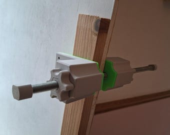 May include: A white and green clamp is attached to a wooden frame. The clamp has a threaded rod and a knob for tightening. The wooden frame is light brown. The clamp is designed to hold objects in place.