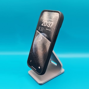 Puede incluir: Un smartphone con funda negra se muestra en un soporte gris para teléfono sobre un fondo azul. La pantalla del teléfono muestra la hora 20:37. El soporte tiene una base rectangular y un soporte curvo.