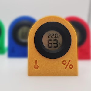 May include: A mustard-yellow digital thermometer and hygrometer showing 22.0°C and 63%. The device has a black circular display and a black ring around it. A thermometer icon and a percentage symbol are on the front.
