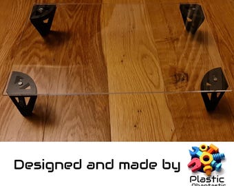 May include: A clear acrylic sheet with four black plastic corner brackets. The sheet is resting on a wooden surface. The text "Designed and made by Plastic Phantastic" is visible below the sheet.