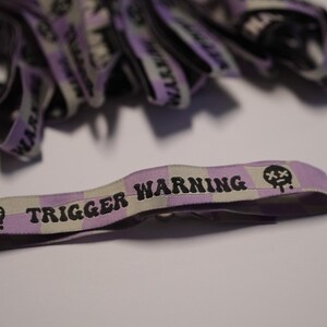 Könnte beinhalten: Nahaufnahme mehrerer Stoffarmbänder. Das primäre Armband ist helllila und weiß kariert mit den Worten "TRIGGER WARNING" in Schwarz. Eine kleine Grafik eines Schädels mit tropfenden Tränen befindet sich an jedem Ende.