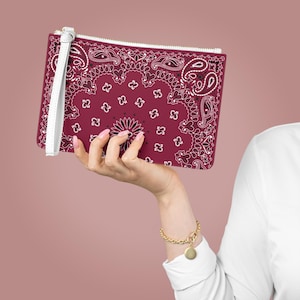 Puede incluir: Un bolso de mano burdeos con cremallera y correa de muñeca blancas. El bolso presenta un estampado de cachemira en blanco y negro. La correa de muñeca es blanca y la cremallera es plateada. Una persona sostiene el bolso.