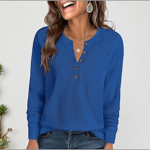 May include: A long-sleeved, royal blue waffle-knit shirt with a Henley neckline. The shirt features a series of four brown buttons down the front and a ribbed cuff at the wrist. The shirt is paired with blue jeans.
