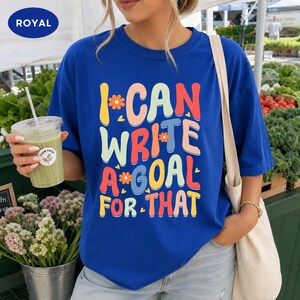 May include: A royal blue short-sleeve t-shirt featuring the colorful text I CAN WRITE A GOAL FOR THAT in a retro font with small flower and heart accents. The shirt has a crew neckline and is worn with a light-colored shoulder bag.