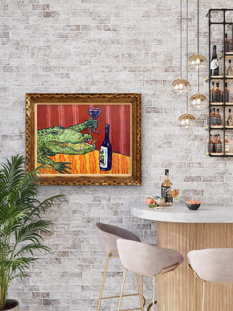 May include: A framed painting featuring a green alligator holding a wine glass filled with red liquid, positioned next to a blue wine bottle with a grape label. The scene is set against a red striped background and an orange patterned tablecloth.
