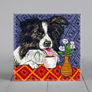 May include: A black and white border collie dog with a pink tongue sticking out is sitting at a table with a cup of coffee. The dog is looking at the camera. The table has a red and orange checkered tablecloth. There is a vase with purple flowers on the table.