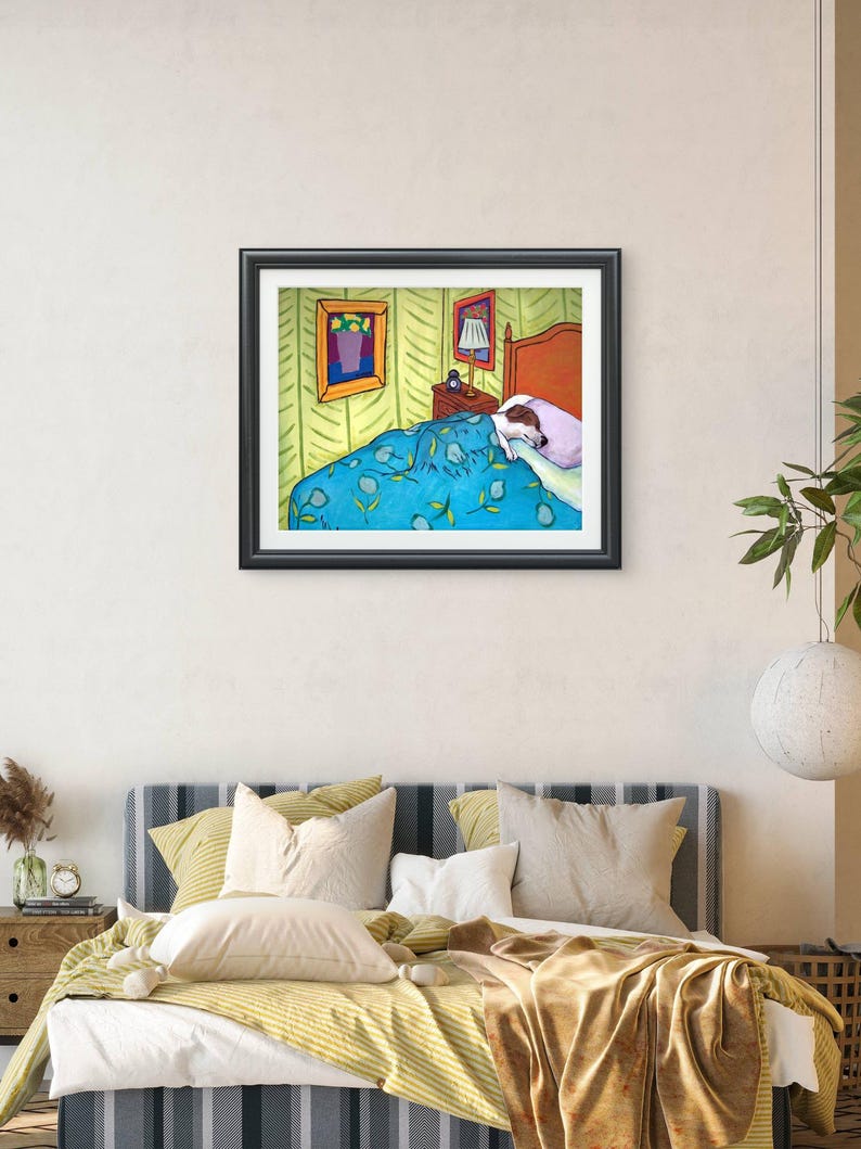 May include: A framed art print depicting a dog sleeping in a bed under a blue blanket with yellow leaf patterns. The bedroom scene includes a bedside table with a lamp, a light-coloured wall, and two framed pictures hanging on the wall above the bed.