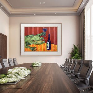May include: A framed wall art print featuring a green alligator sitting at a table with an orange patterned tablecloth. The alligator holds a glass of red wine, and a blue wine bottle with a grape label sits on the table against a red striped background.
