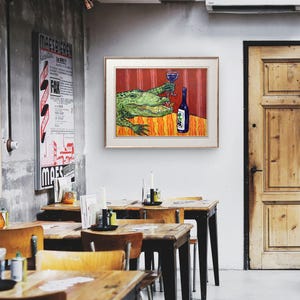 May include: A framed wall art print featuring a green alligator sitting at a table with a blue wine glass and a bottle of wine. The background is a vibrant red with vertical stripes, and the table has an orange and yellow patterned surface.