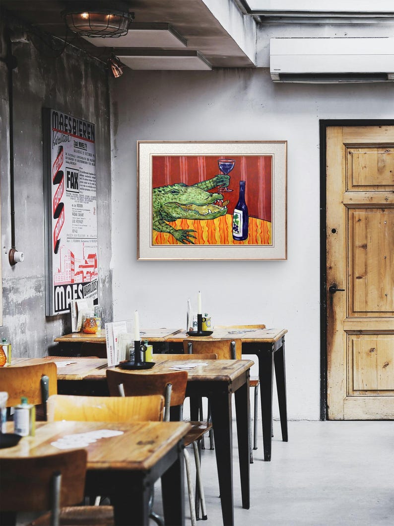 May include: A framed wall art print featuring a green alligator sitting at a table with a blue wine glass and a bottle of wine. The background is a vibrant red with vertical stripes, and the table has an orange and yellow patterned surface.