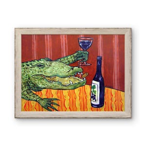 May include: A framed painting depicts a green alligator with an open mouth holding a wine glass in one claw. A wine bottle with a grape label sits on an orange patterned tablecloth against a red striped background. The artist signature is visible.