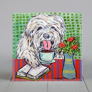Puede incluir: Un Old English Sheepdog blanco con la lengua rosa que sobresale está bebiendo café de una taza azul y blanca. El perro está sentado en una mesa con un mantel rojo y morado y un libro. Hay flores rojas en un jarrón azul y amarillo sobre la mesa.