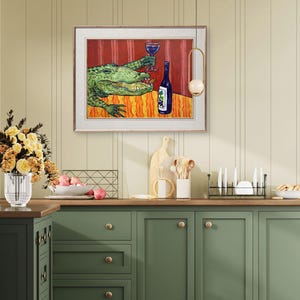 May include: A framed wall art print featuring a green alligator sitting at a table with a red tablecloth, holding a glass of red wine in one claw and a blue wine bottle with a grape label sitting on the table next to it, set in a kitchen.