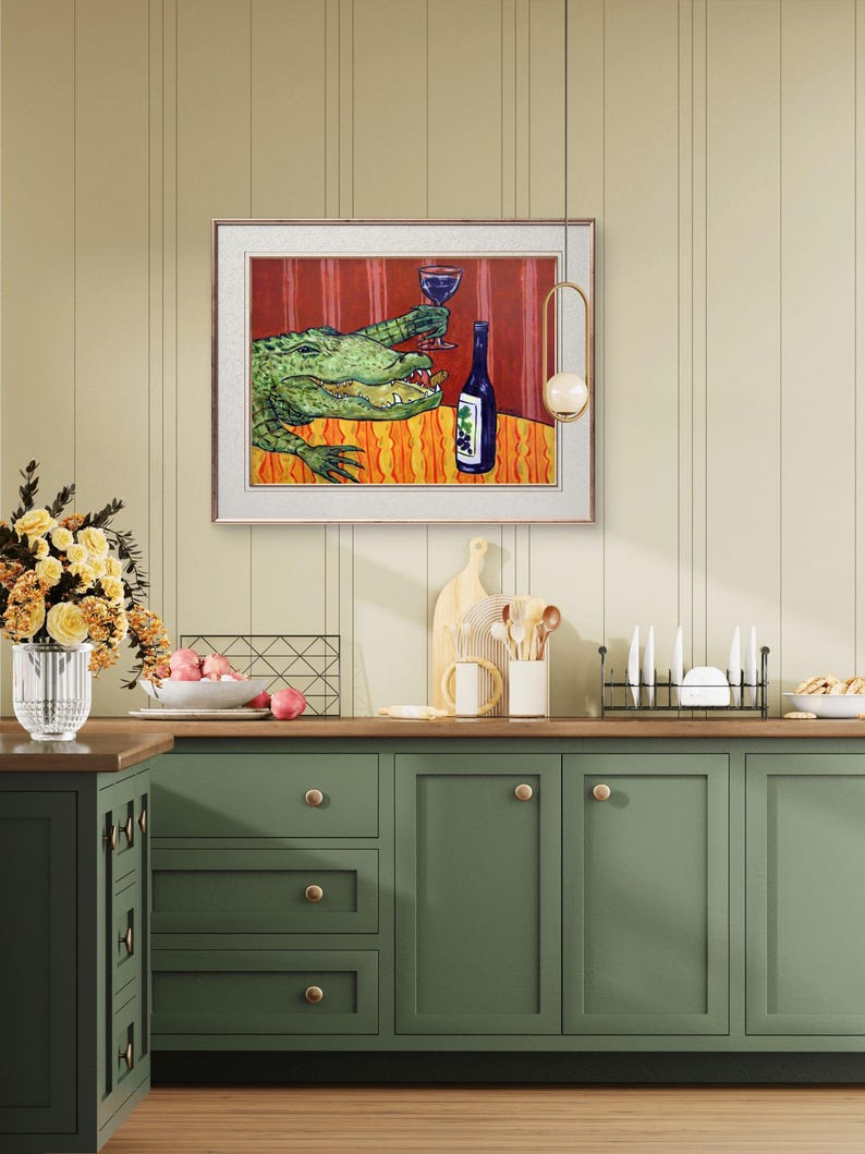May include: A framed wall art print featuring a green alligator sitting at a table with a red tablecloth, holding a glass of red wine in one claw and a blue wine bottle with a grape label sitting on the table next to it, set in a kitchen.
