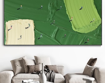 May include: Abstract golf course painting in a black frame. The artwork shows a textured green and light yellow surface with miniature figures of golfers. The painting is displayed above a modern living room sofa.