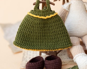 May include: A handmade crochet doll with a green dress, brown shoes, and a sunflower-themed blanket. The dress is olive green with yellow trim. The doll has brown hair and is wearing a white dress.