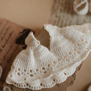 May include: A white crocheted baby dress with a ruffled hem and a pearl button detail.